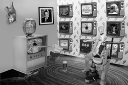 A surreal image. The scene is a living room with one large vintage TV and a wall of nine other smaller TVs. There is a Luminary sitting in a vintage dentist chair with his hand out. Inside the large TV is a Luminary character with a lighthouse for a head and a Pinocchio nose. He is pouring Kool-Aid from a vintage Kool-Aid Picther into a glass on the floor. The other TVs all have symbols of something disingenuous and all TVs have logo of a head of a fox. There is a opossum hanging from the ceiling and an alligator on the floor. There is a portrait of Nixon on the wall.