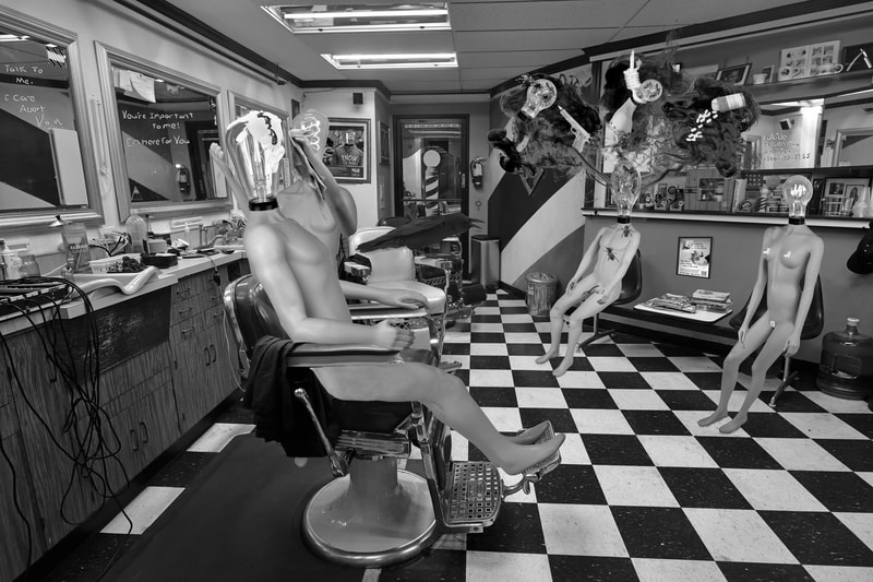 A surreal image. The scene is in a vintage looking barber shop. There is a Luminary barber who instead of cutting hair is wiping bird poop off of the client Luminary's light bulb. On the other side of the room a young male Luminary with a female light bulb has black smokey looking thoughts coming from his light bulb. The smoke is showing three dark thoughts. One shows a handgun shooting his head. One shows his neck in a hangman's noose and the last shows a bottle of pills with the pills spilling out. An older female Lumarian sits next to him. There are mirrors all around the barber shop and each one has writing on them one is the suicide hotline phone number.  