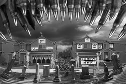 A surreal composite photo.  There are two houses side by side. One house has shooting targets, classic pro gun posters on it and the other has Screw the NRA on it. There are two female Luminaries building a wall between the two houses. There is a male luminary with an assault rifle and one with a large pen. In the street there are grave stones and overhead there are 223 bullets. They form a sharks mouth. 